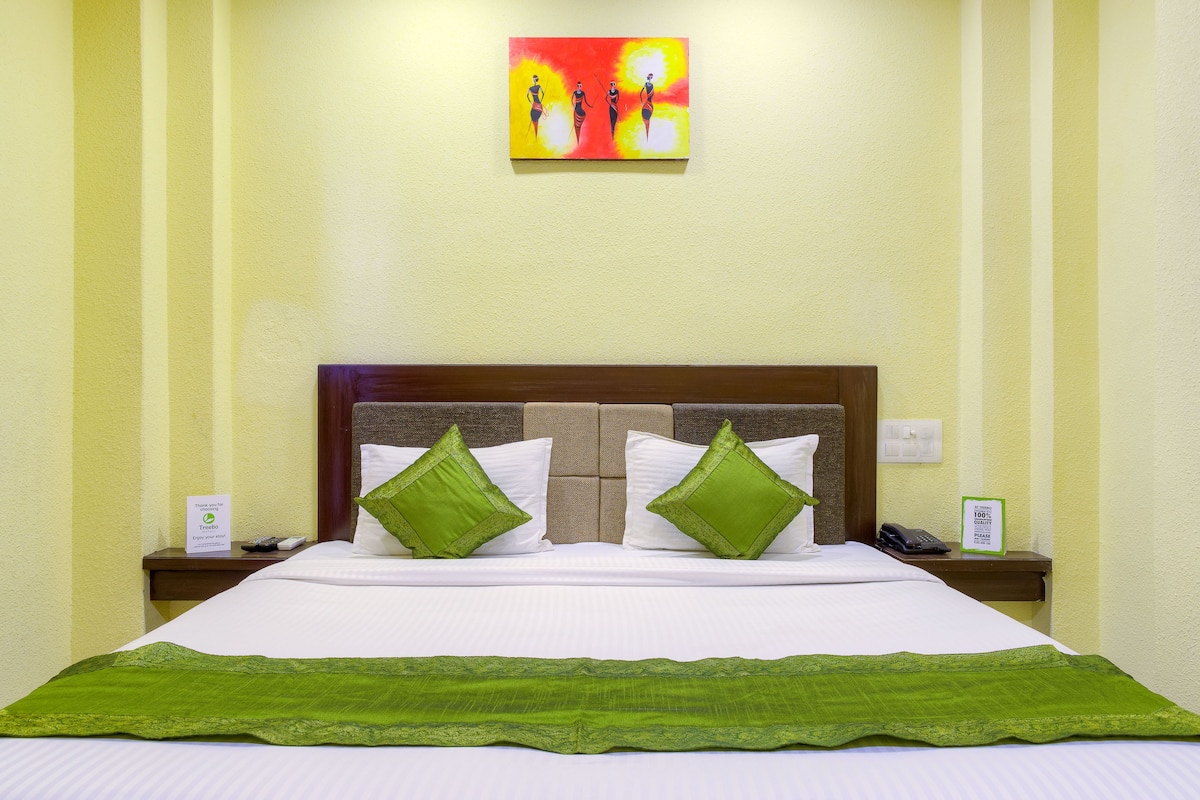 Habitación de hotel con cama, almohadas verdes, cabecero, mesita de noche con teléfono y un cuadro en la pared, además de un cartel en la mesita.