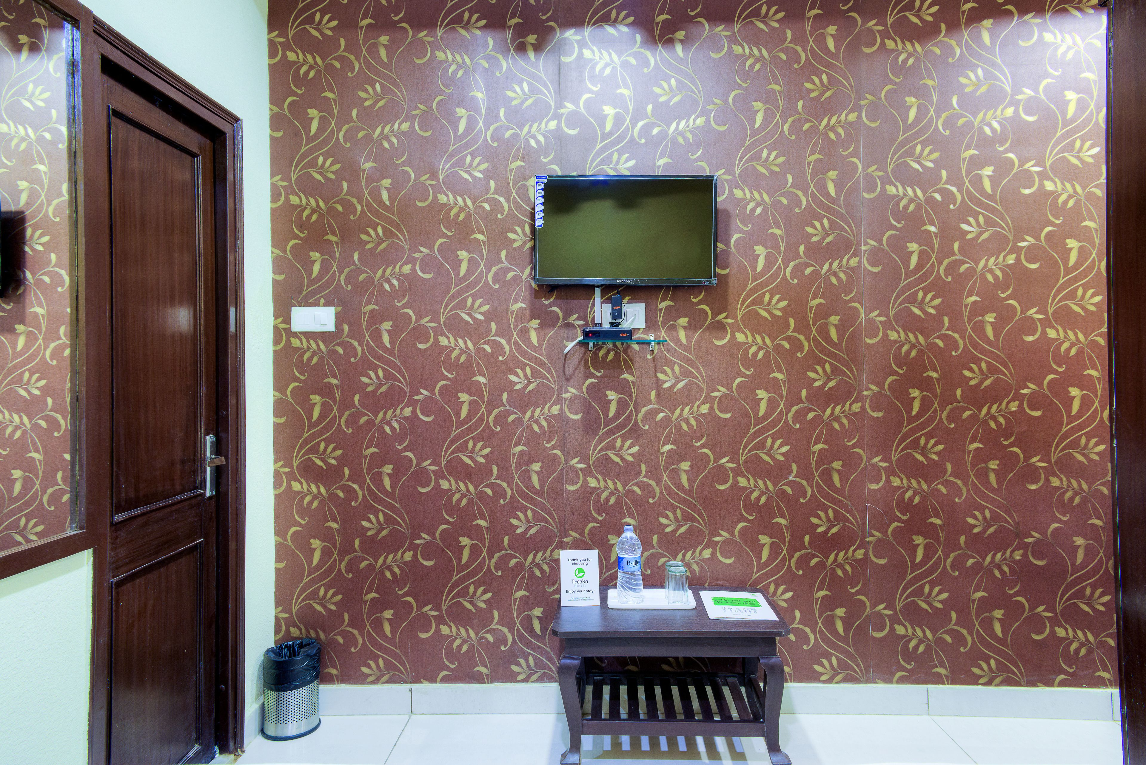 Habitación de hotel con una televisión montada en la pared, una mesita con una botella de agua y un vaso, y un cubo de basura.
