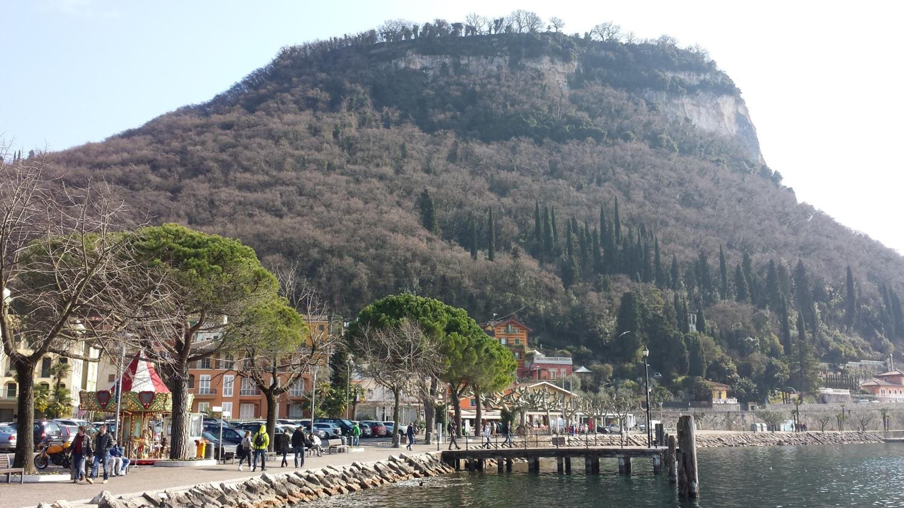 A waterfront promenade with a mountain backdrop, featuring a carousel, parked cars, and a pier extending into the water.