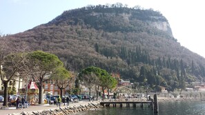 Un paseo marítimo con montañas al fondo, que incluye un carrusel, coches aparcados y un muelle que se adentra en el agua.