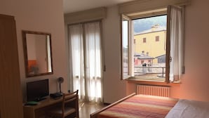 Un dormitorio con cama, escritorio con computadora, silla, ventana con vista a edificios y un radiador.
