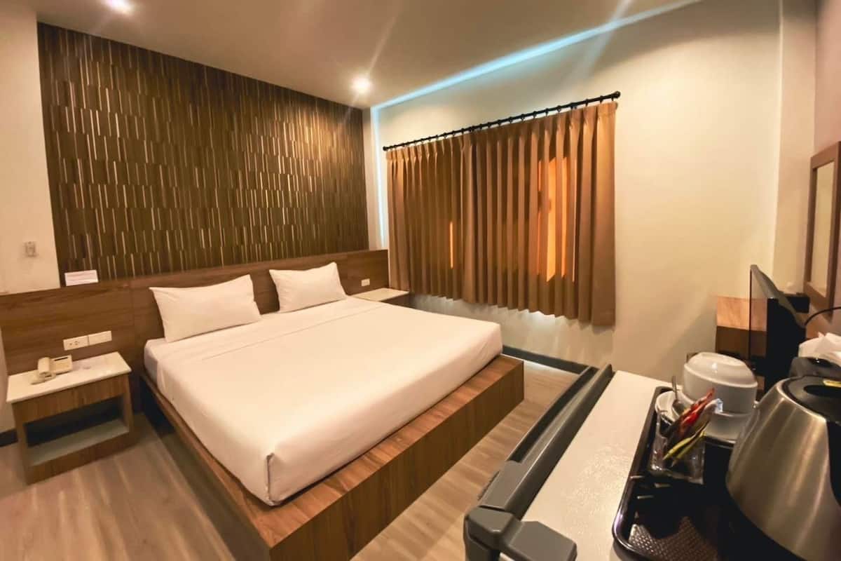 A modern hotel room with a large bed, wooden headboard, a bedside table, a desk with a chair, and a coffee maker.