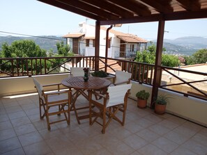 A covered outdoor patio with a round wooden table and chairs, potted plants, and a view of a hillside town. - Villa Helidoni (Platanias)