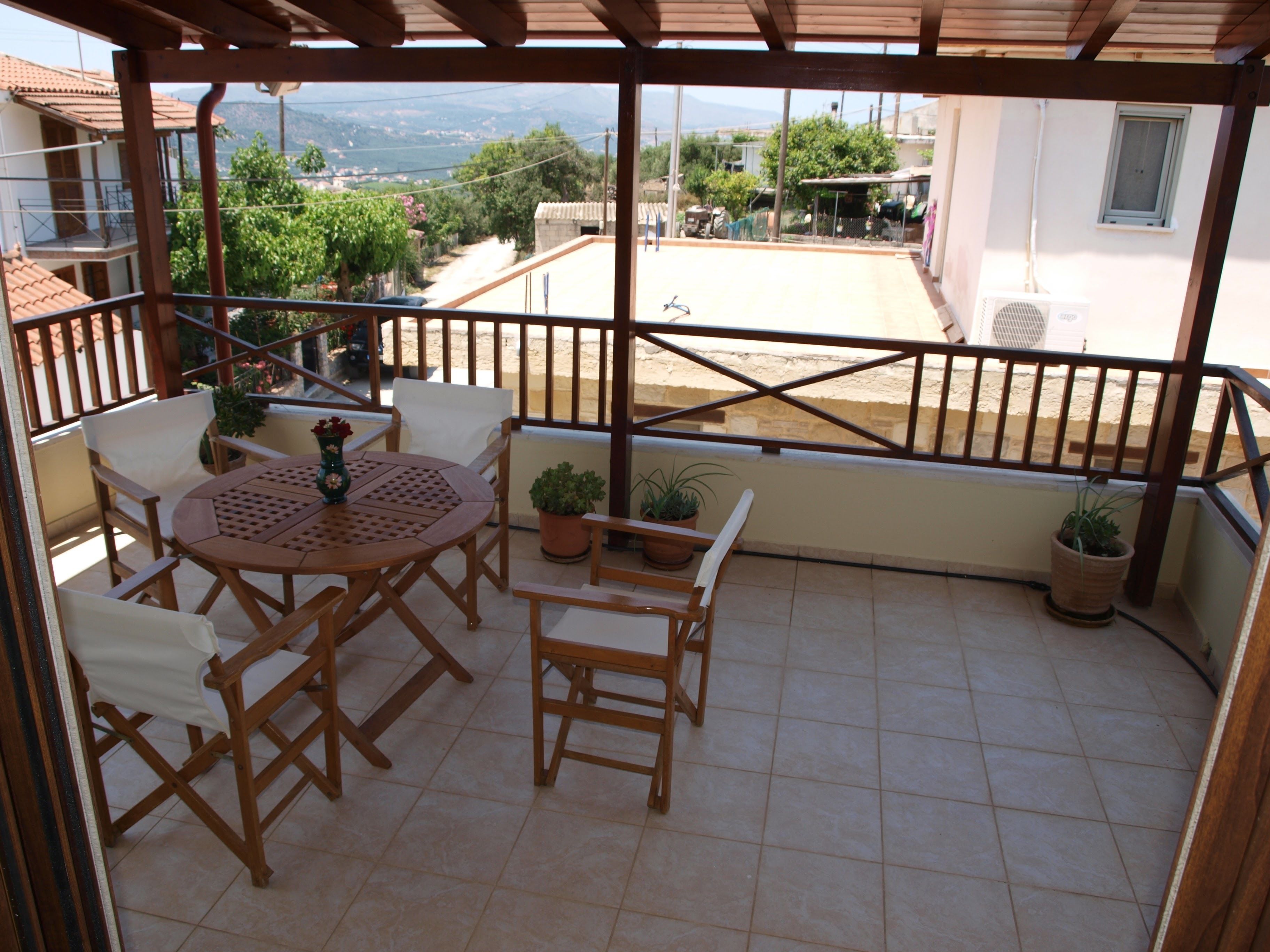 Eine überdachte Terrasse mit Holztisch und -stühlen, Blumentöpfen und Blick auf die umliegende Landschaft.