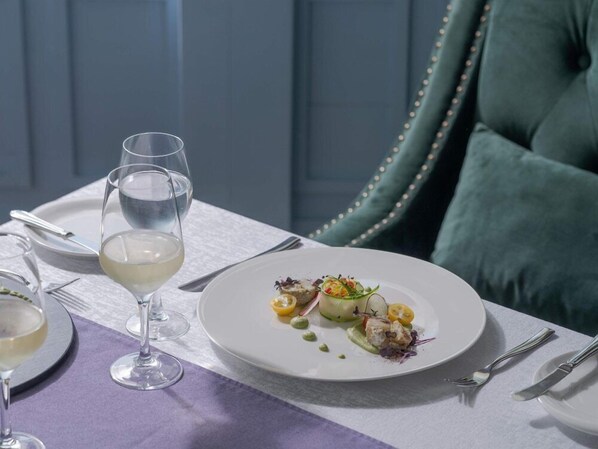 Une table à manger avec une assiette blanche remplie de nourriture, un verre de vin blanc et des couverts en argent.