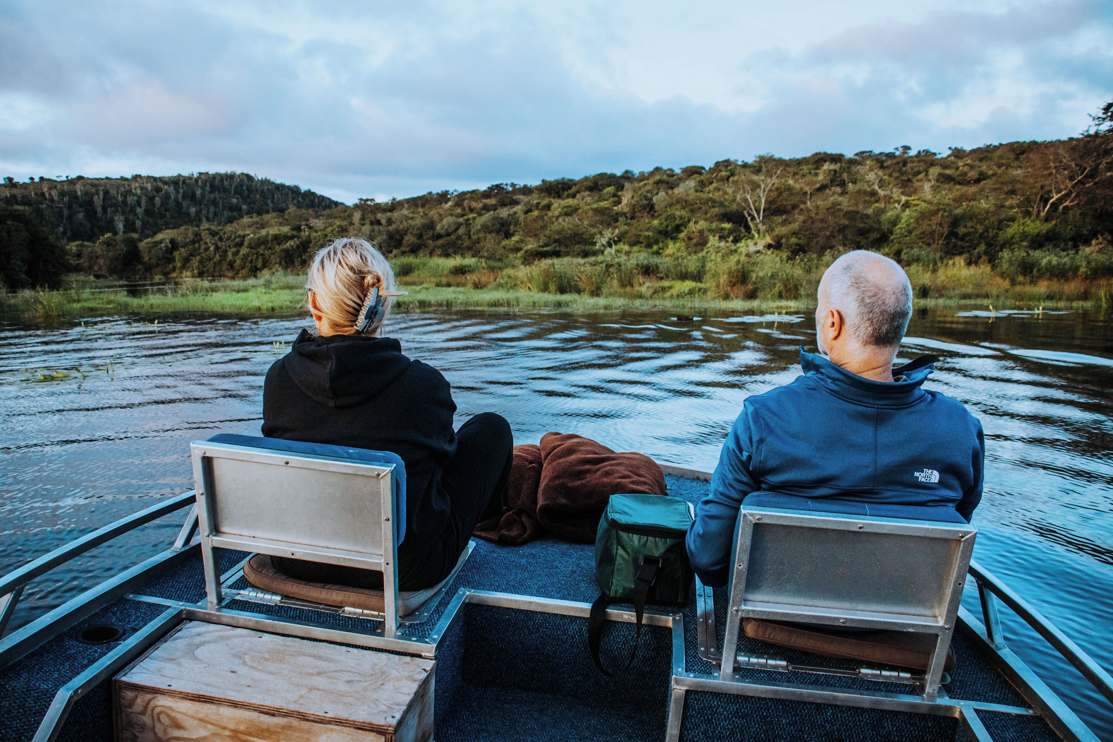 Twee personen zitten op een boot met metalen banken, kijkend uit over een rustige rivier met een bebost heuvellandschap op de achtergrond.
