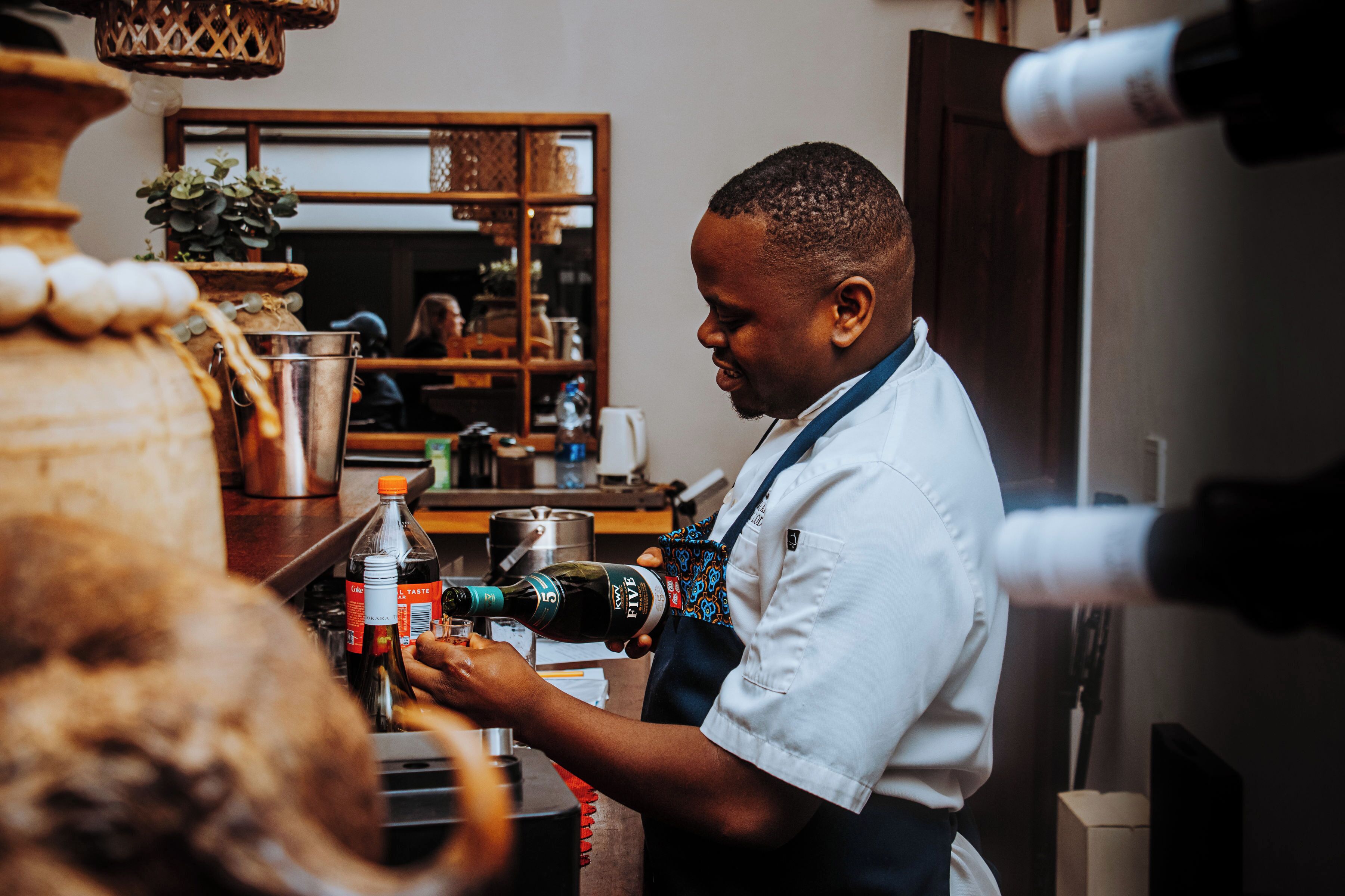 A person in a white shirt and apron preparing a drink at a bar counter with a bottle of Coca-Cola and a bottle of wine.