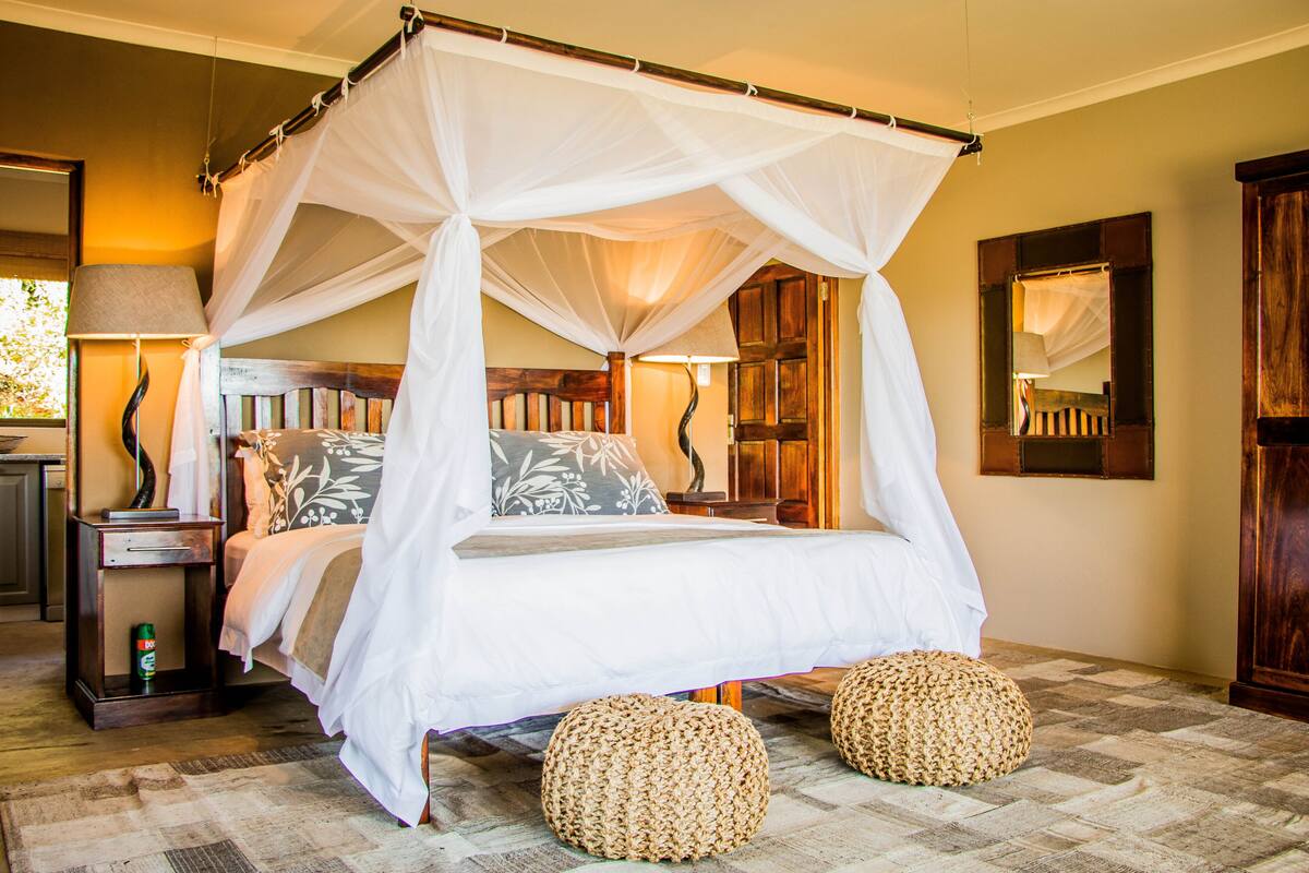 A four-poster canopy bed with a white canopy, two woven ottomans, a nightstand with a lamp, and a mirror on the wall.
