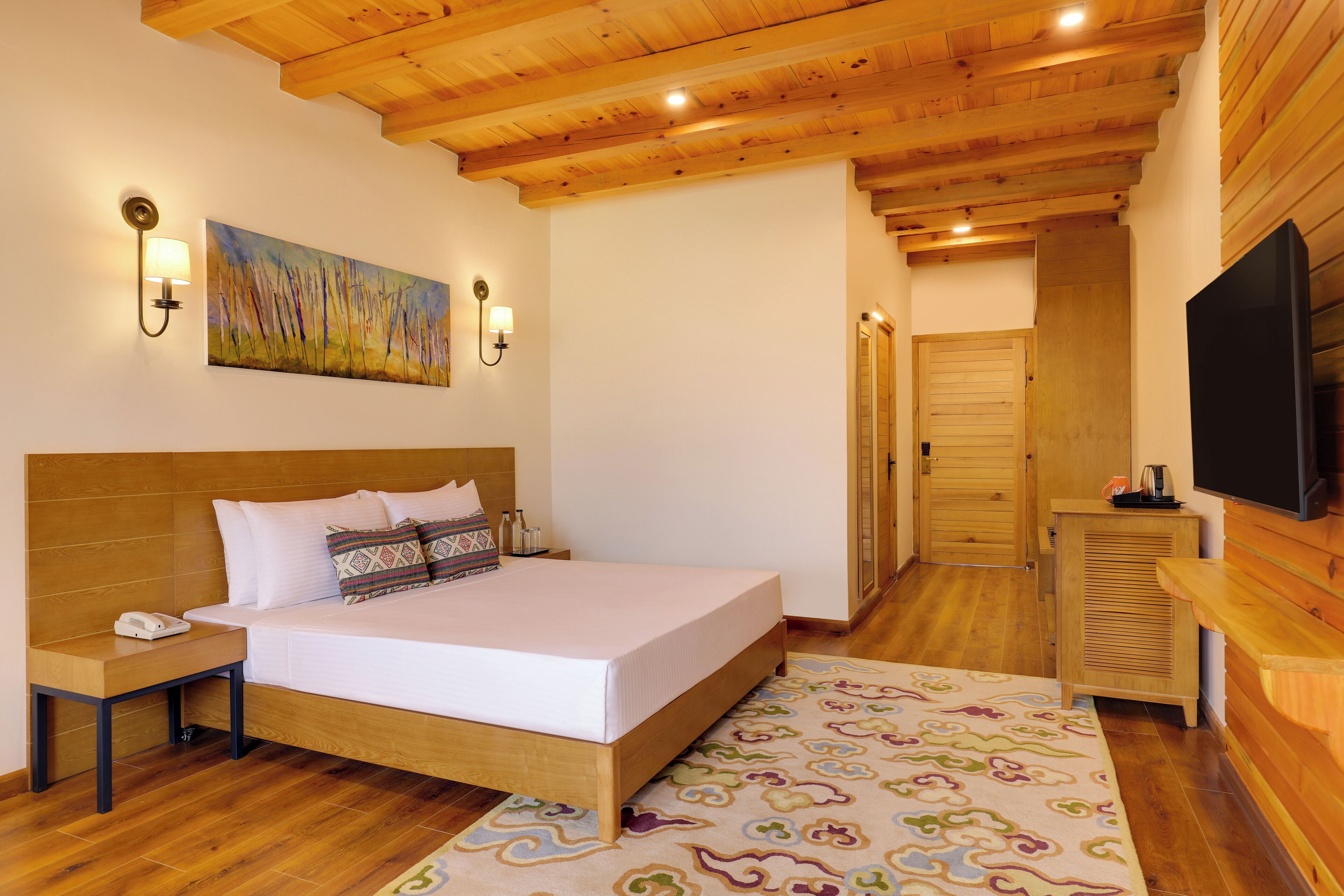 Een slaapkamer met een houten hoofdbord, een bed met witte lakens, een nachtkastje met een telefoon, een wandtelevisie en een schilderij boven het bed.