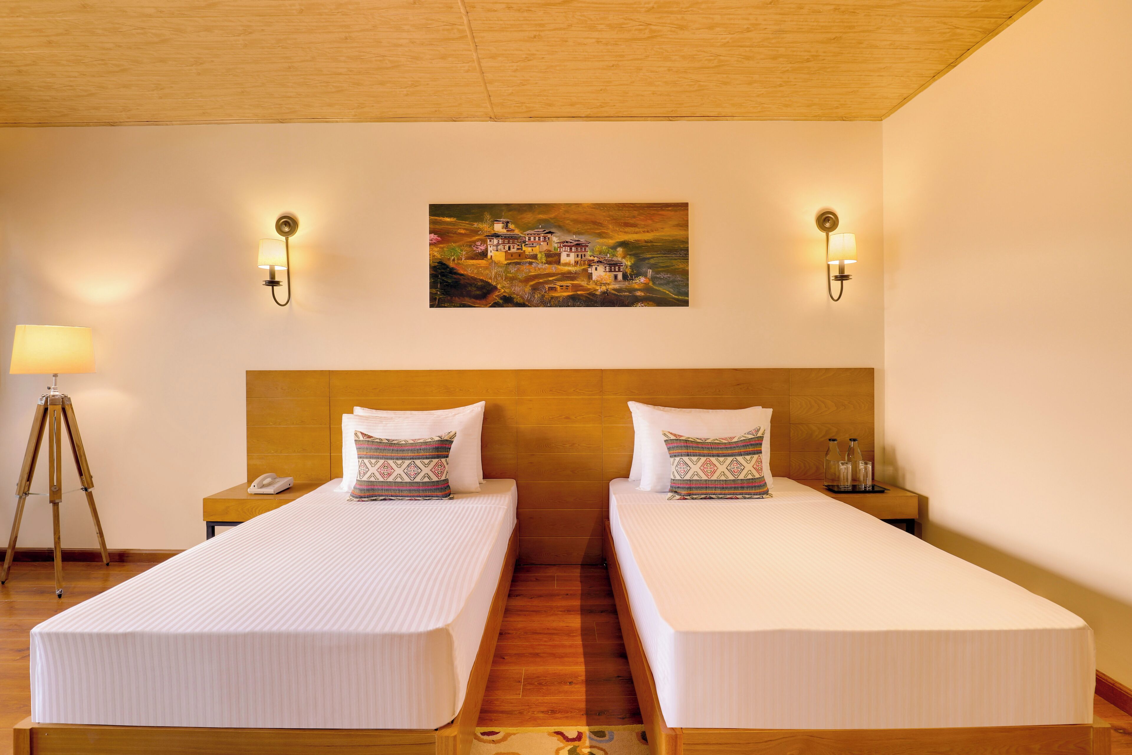 Habitación de hotel con dos camas individuales, cabecero de madera, mesitas de noche con lámparas, un cuadro en la pared y suelo de madera.