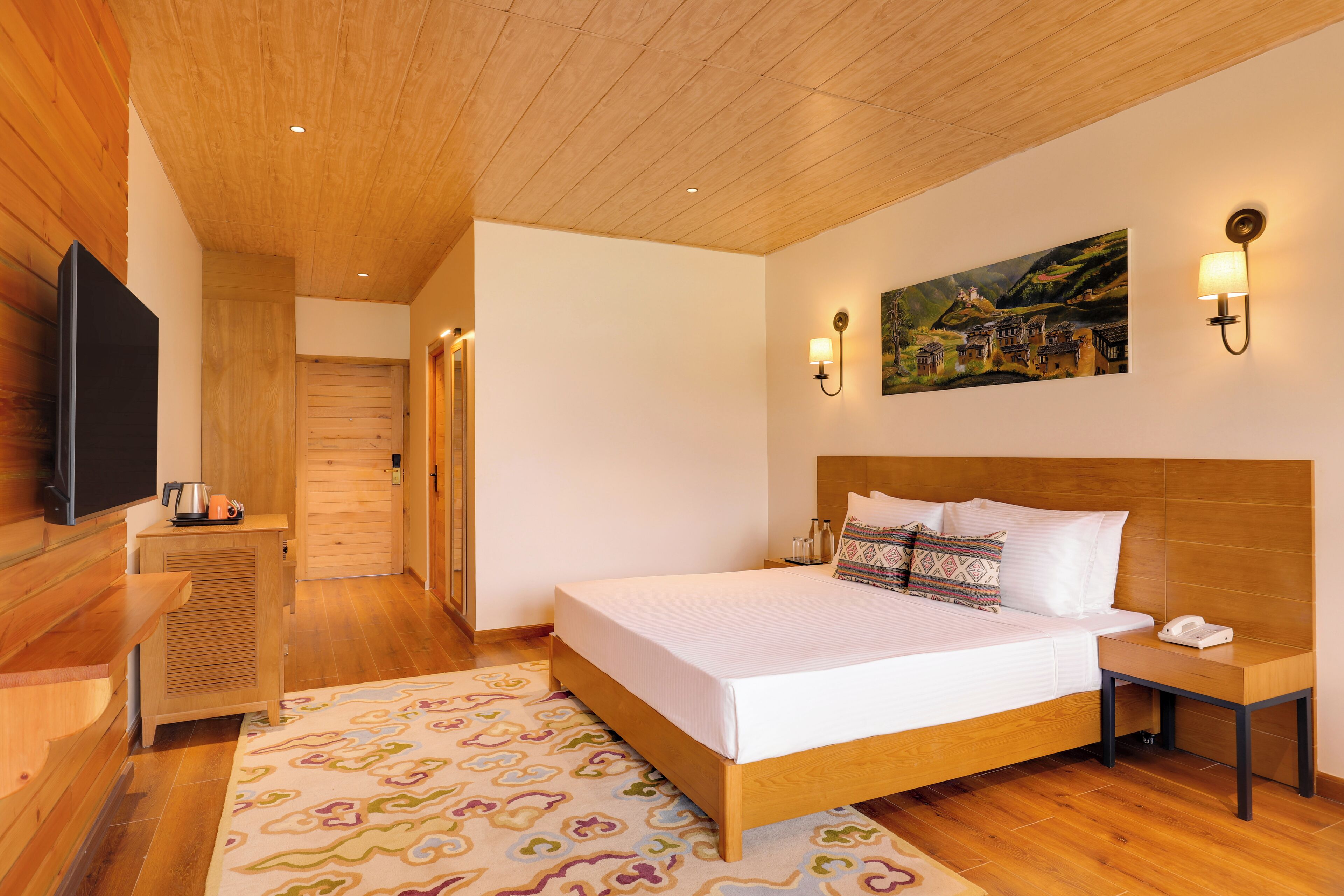 A bedroom with a wooden ceiling, a bed with white linens, a nightstand, a wall-mounted TV, and a painting of a village.