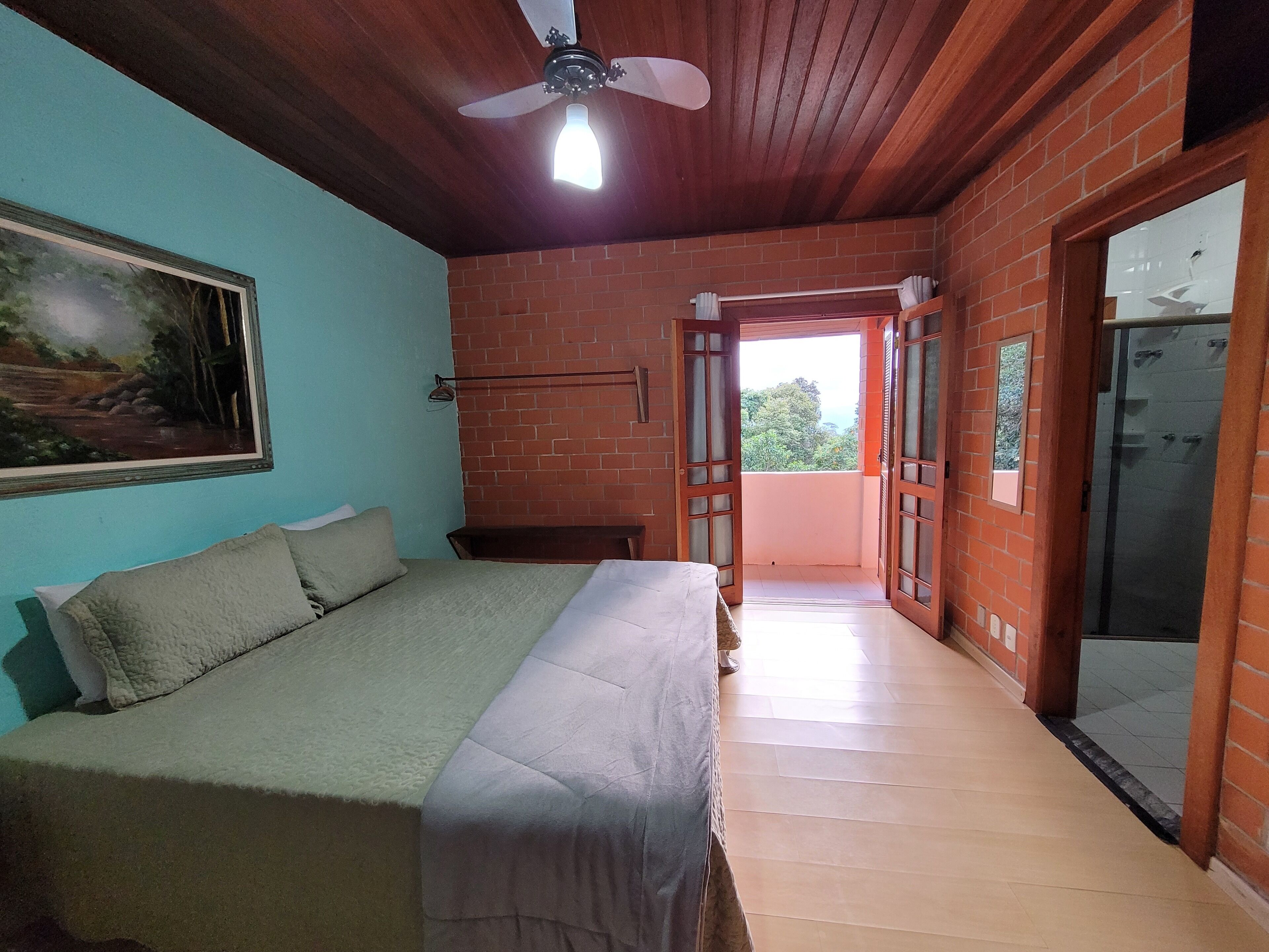 Une chambre à coucher avec un lit, un ventilateur de plafond, un tableau et une vue sur l’extérieur, visible à travers une porte ouverte.
