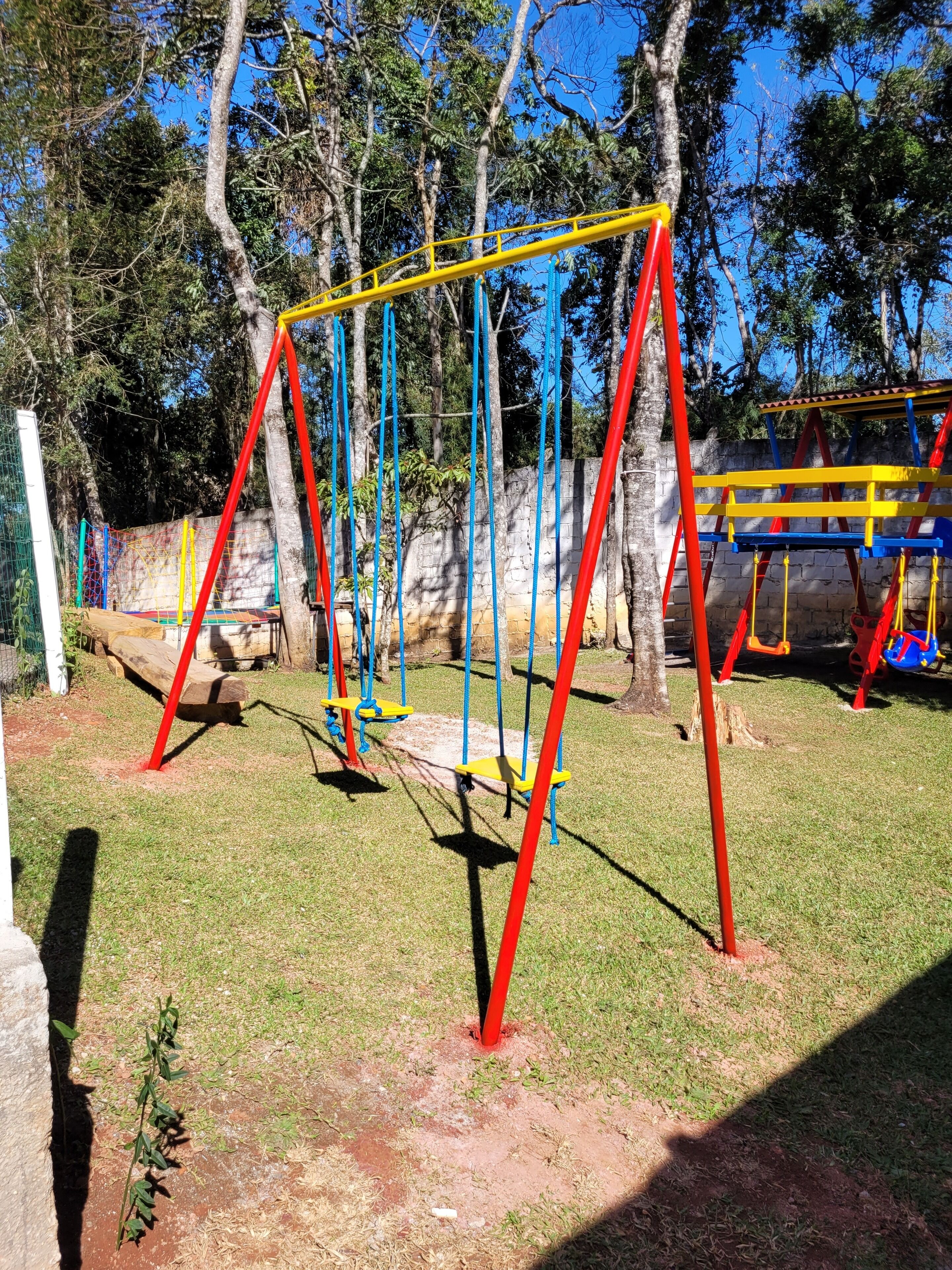 Un terrain de jeux avec des balançoires et des structures d’escalade, entouré d’arbres et délimité par un mur de pierre.