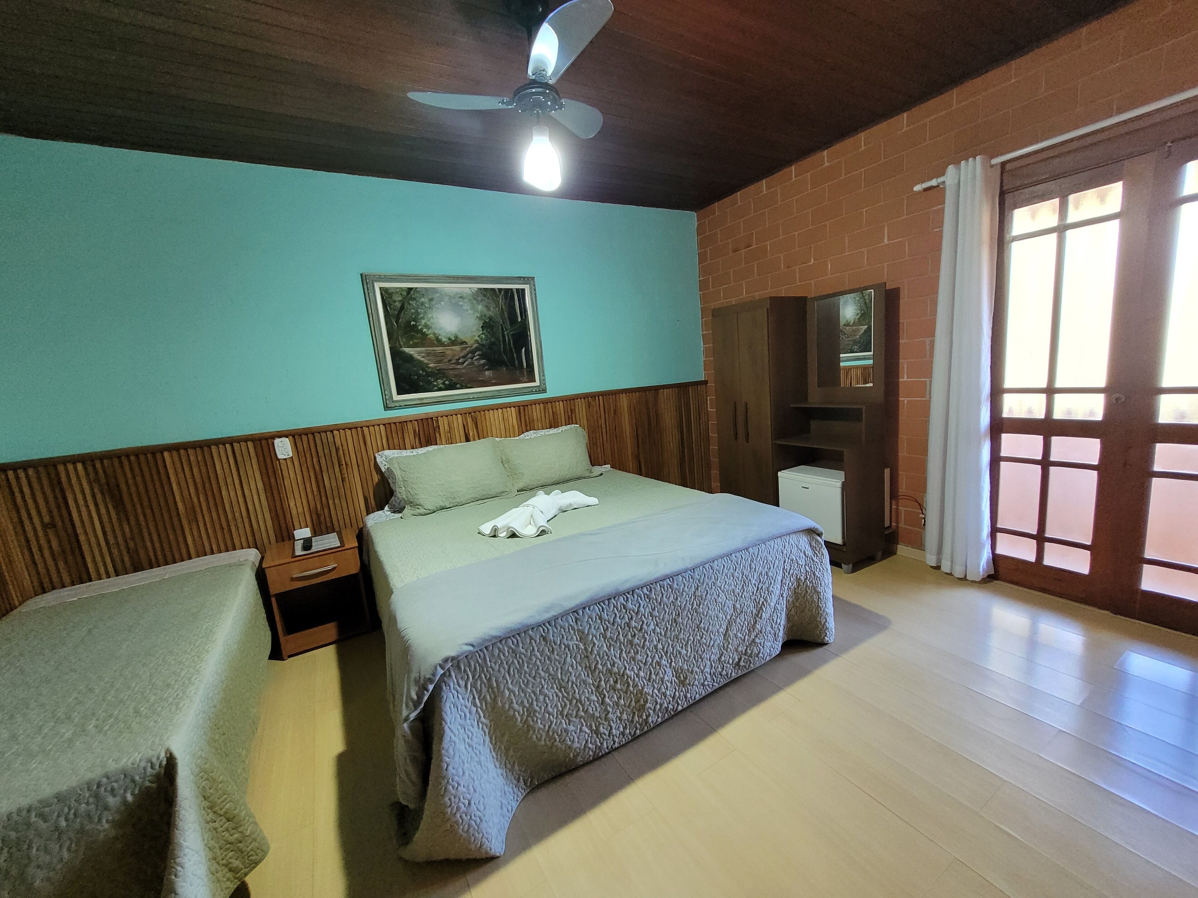 Habitación de hotel con cama, mesita de noche, una pequeña mesa, ventilador de techo, un cuadro en la pared, armario y ventana con cortinas.