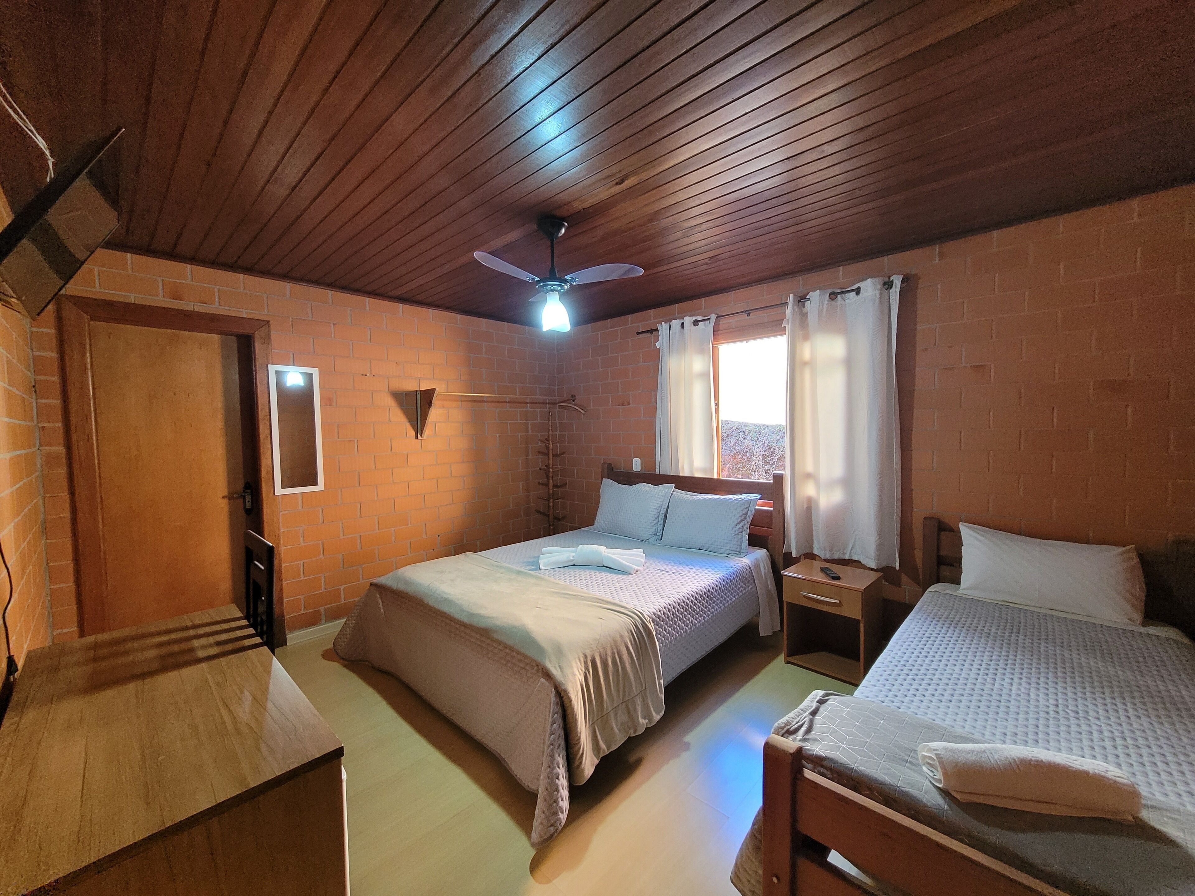 Habitación con dos camas, techo de madera, pared de ladrillo, ventana con cortinas y ventilador de techo.