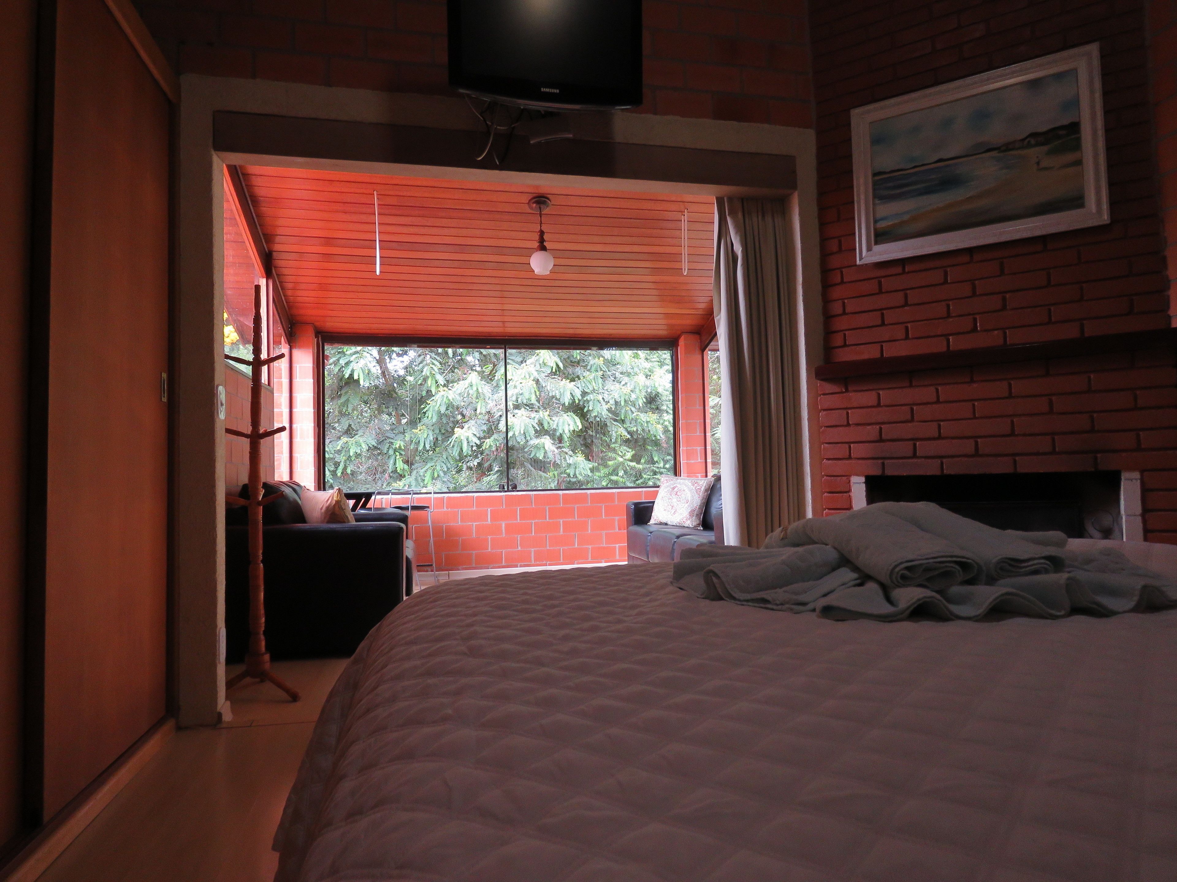 A bedroom with a large bed, a TV mounted on the wall, a window with a view of trees, and a brick fireplace.