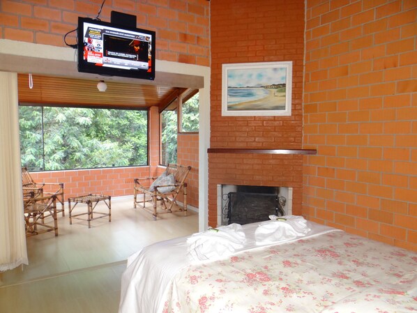 Un dormitorio con pared de ladrillo, un televisor sobre la chimenea, una cama con colcha estampada y vistas a la vegetación a través de grandes ventanales.