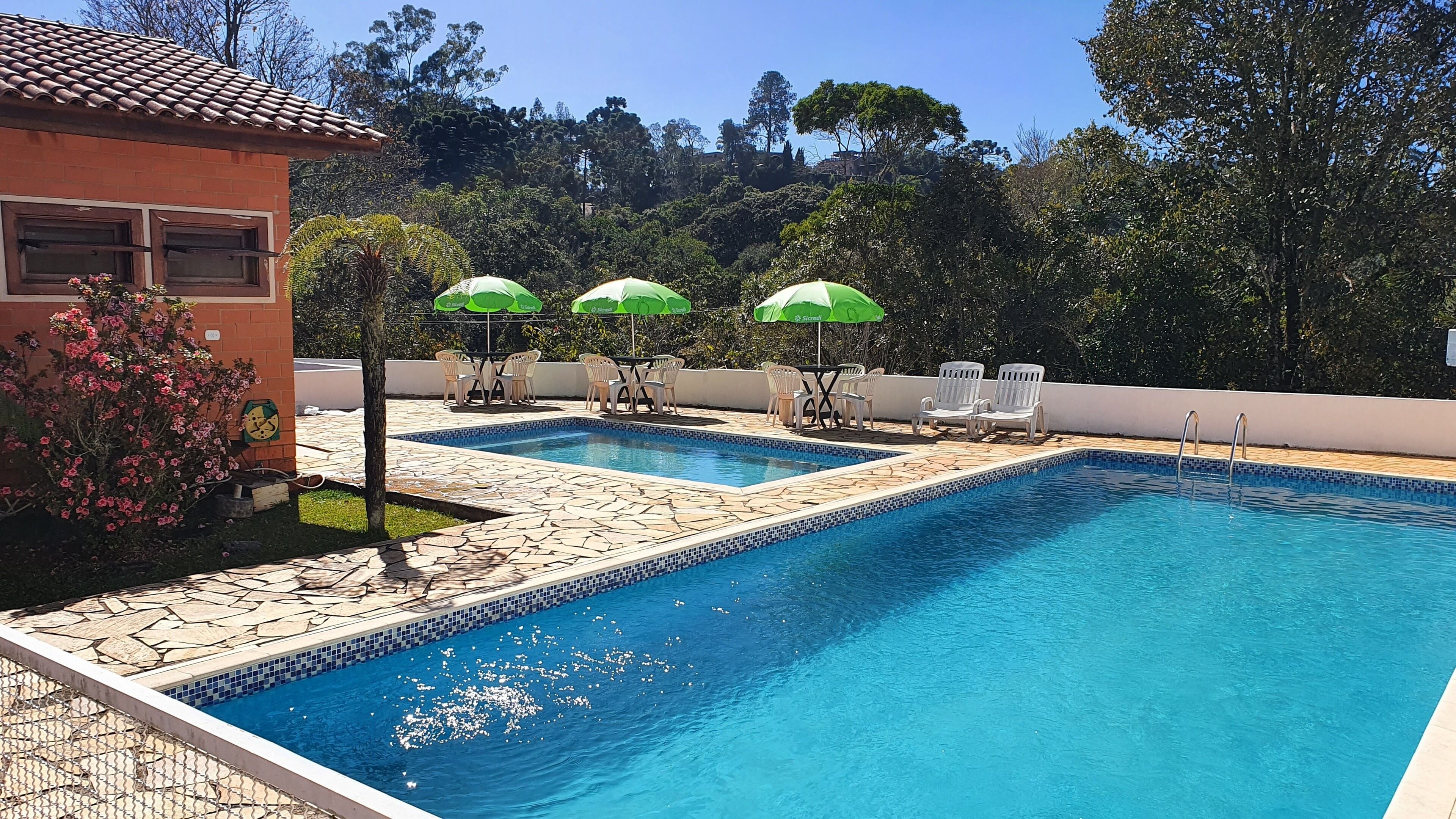 Une piscine rectangulaire, bordée de deux piscines plus petites, entourée de carreaux de pierre et d’une bordure carrelée. On y trouve quatre chaises longues blanches et trois parasols verts.