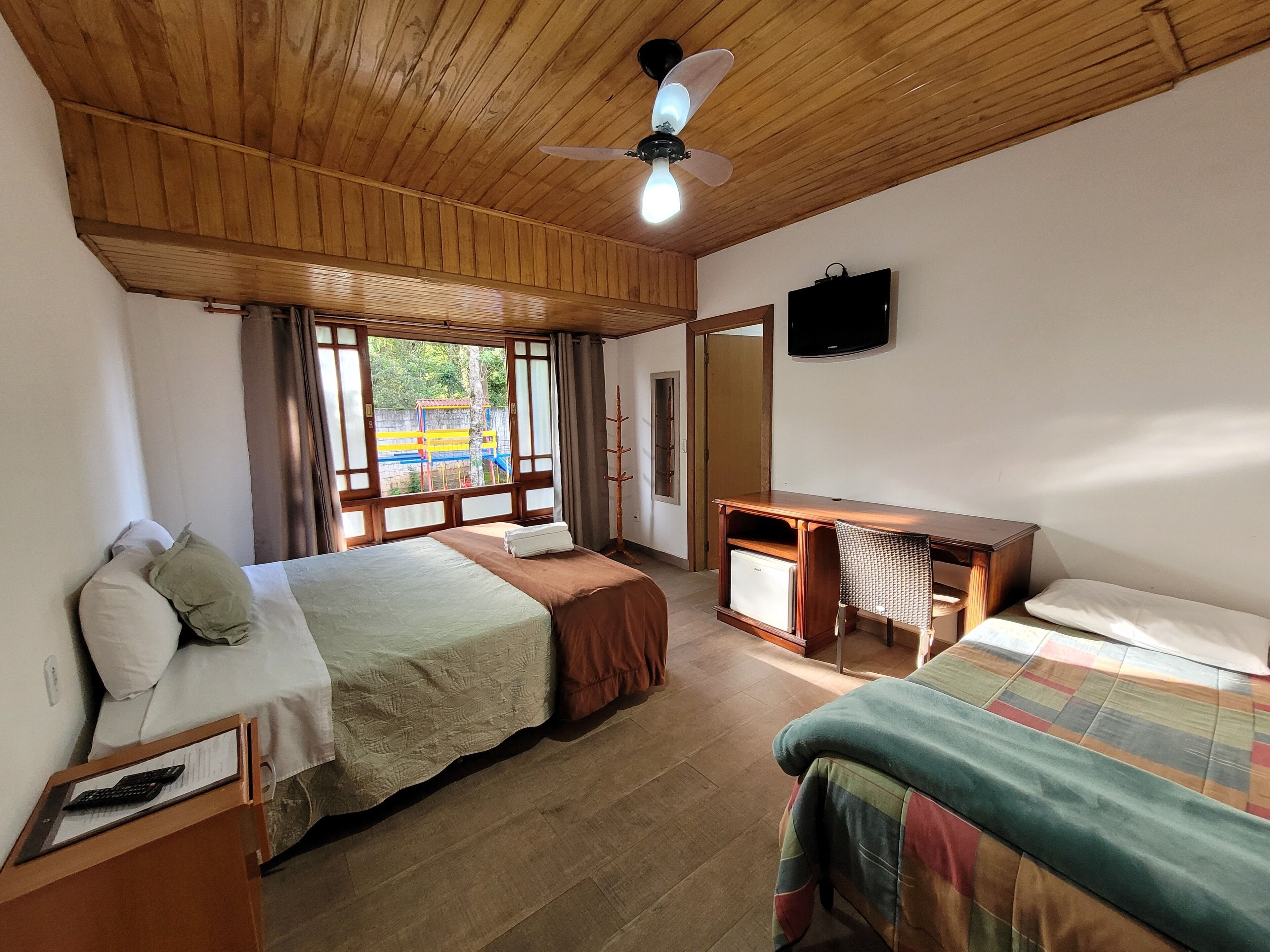 Una habitación con dos camas, un escritorio, un televisor y una ventana con vista a un parque infantil.