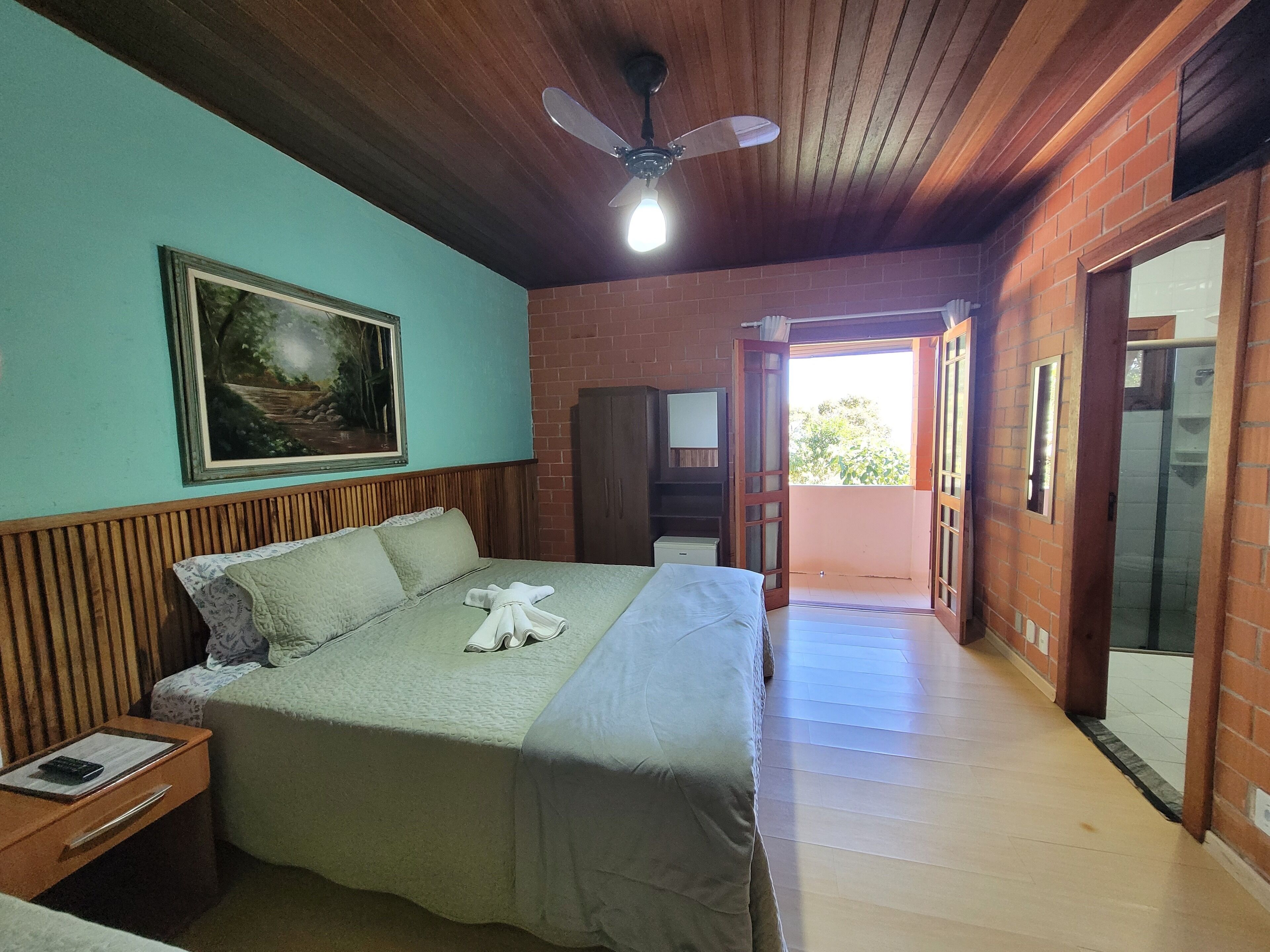 A bedroom with a bed, a nightstand, a painting, a ceiling fan, and a view of the outside.