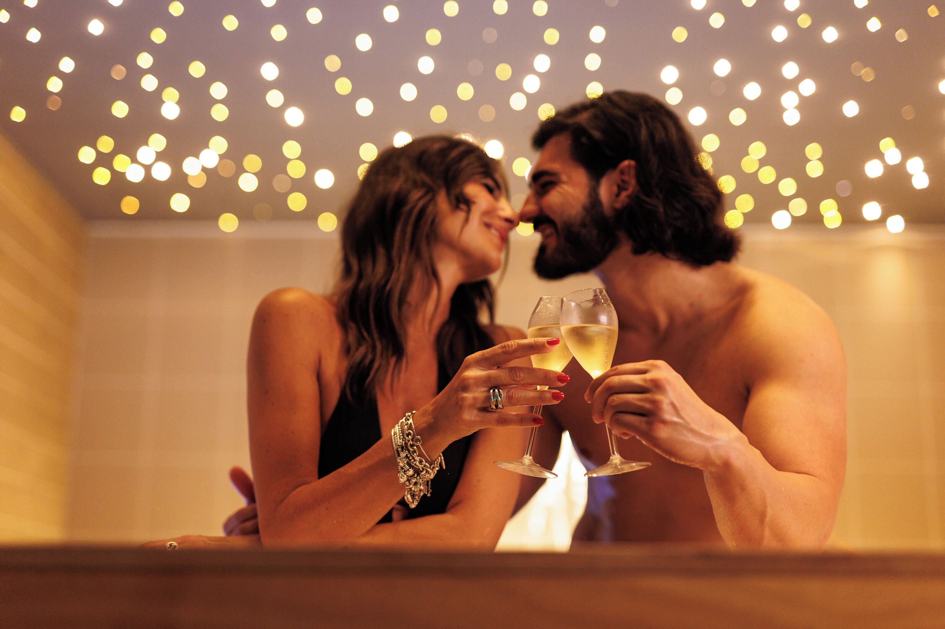 A couple holding champagne glasses in a dimly lit room with string lights in the background.