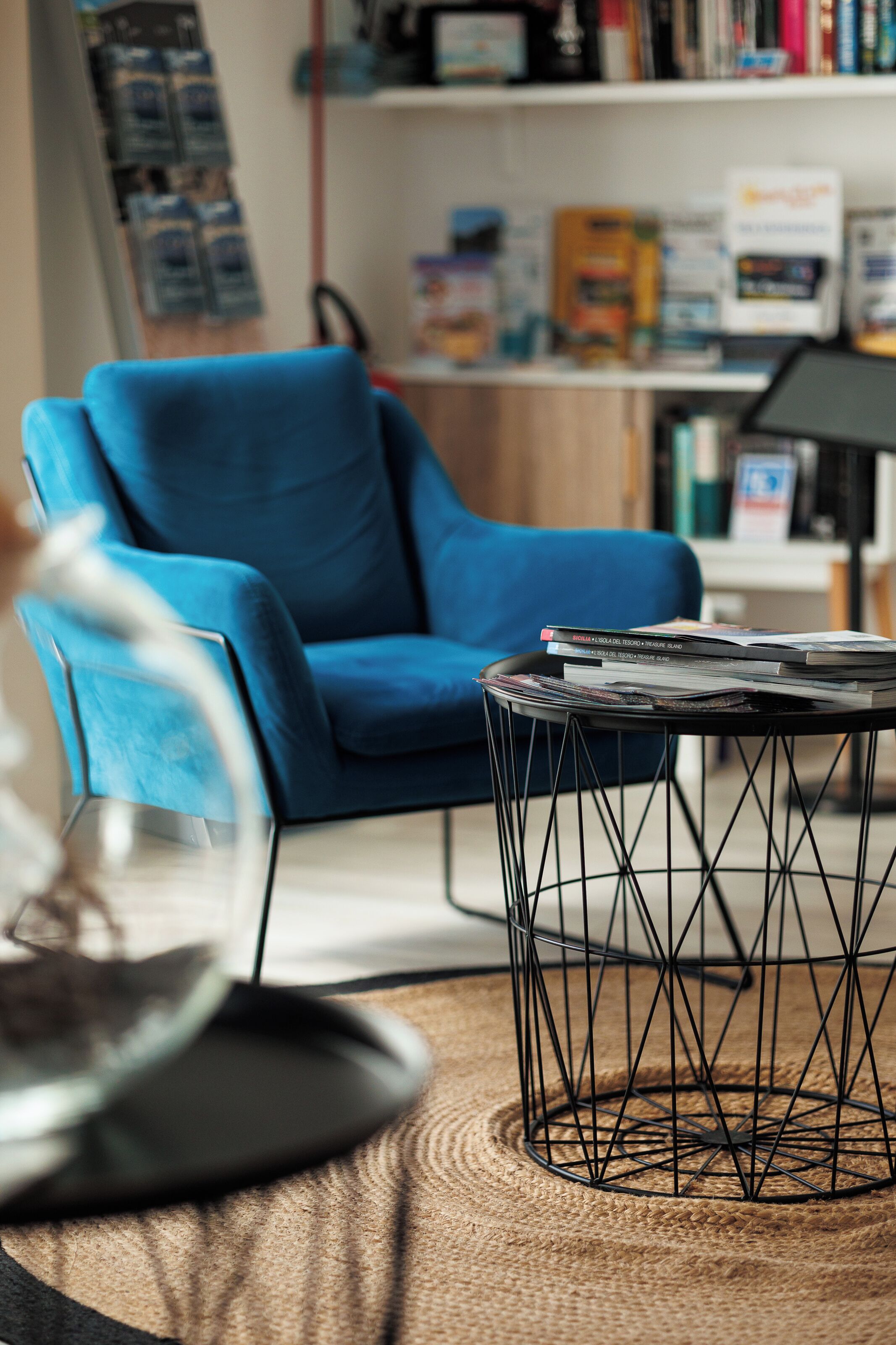 Una habitación con un sillón azul, una mesa de centro redonda con revistas, y una estantería al fondo.