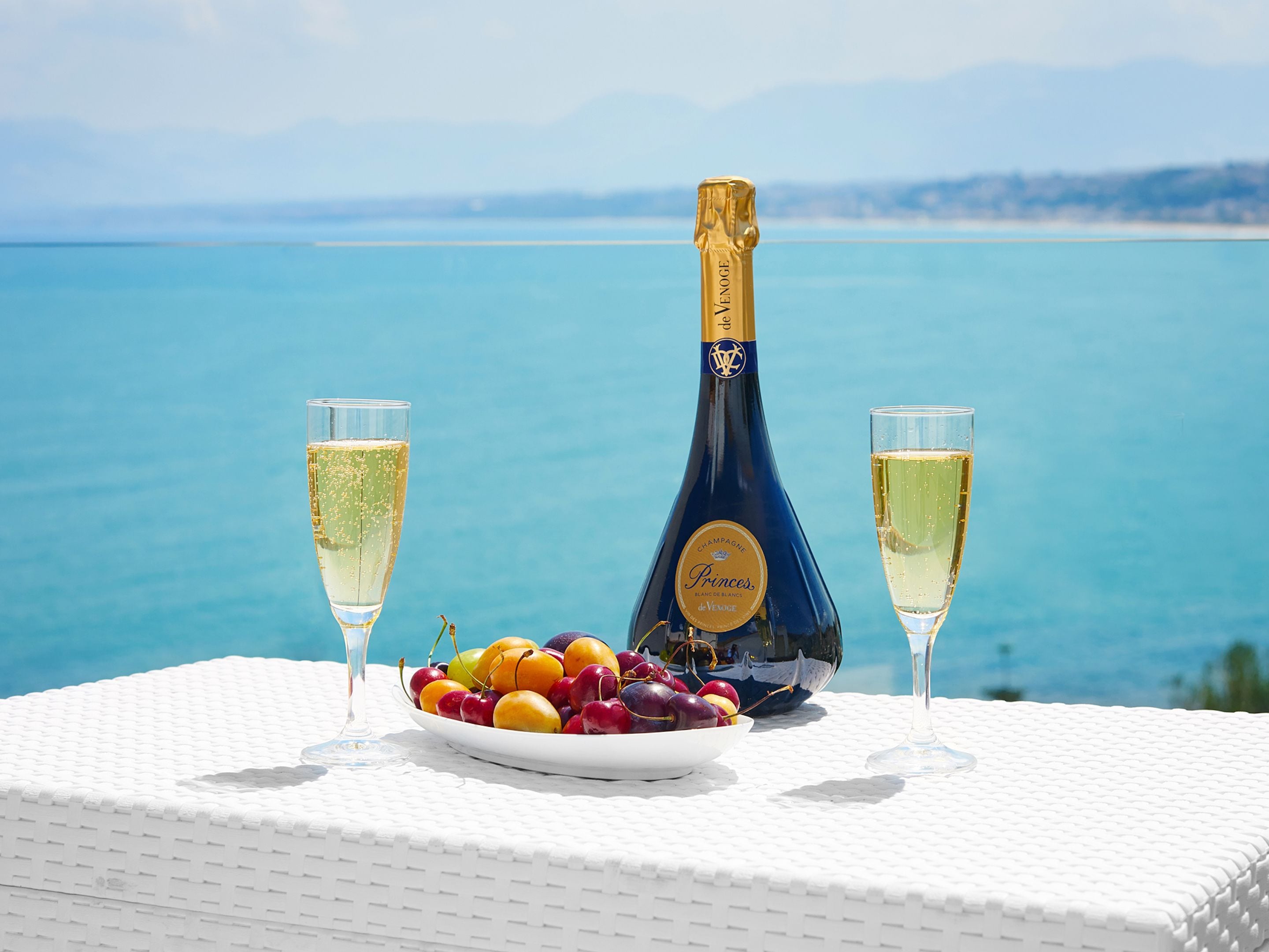 Una botella de champán, dos copas y un bol de fruta sobre una mesa con vistas al mar.