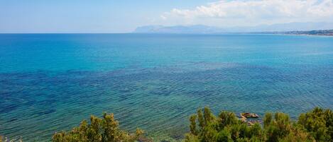 Paisaje costero con mar azul cristalino, montañas a lo lejos y árboles verdes frondosos.
