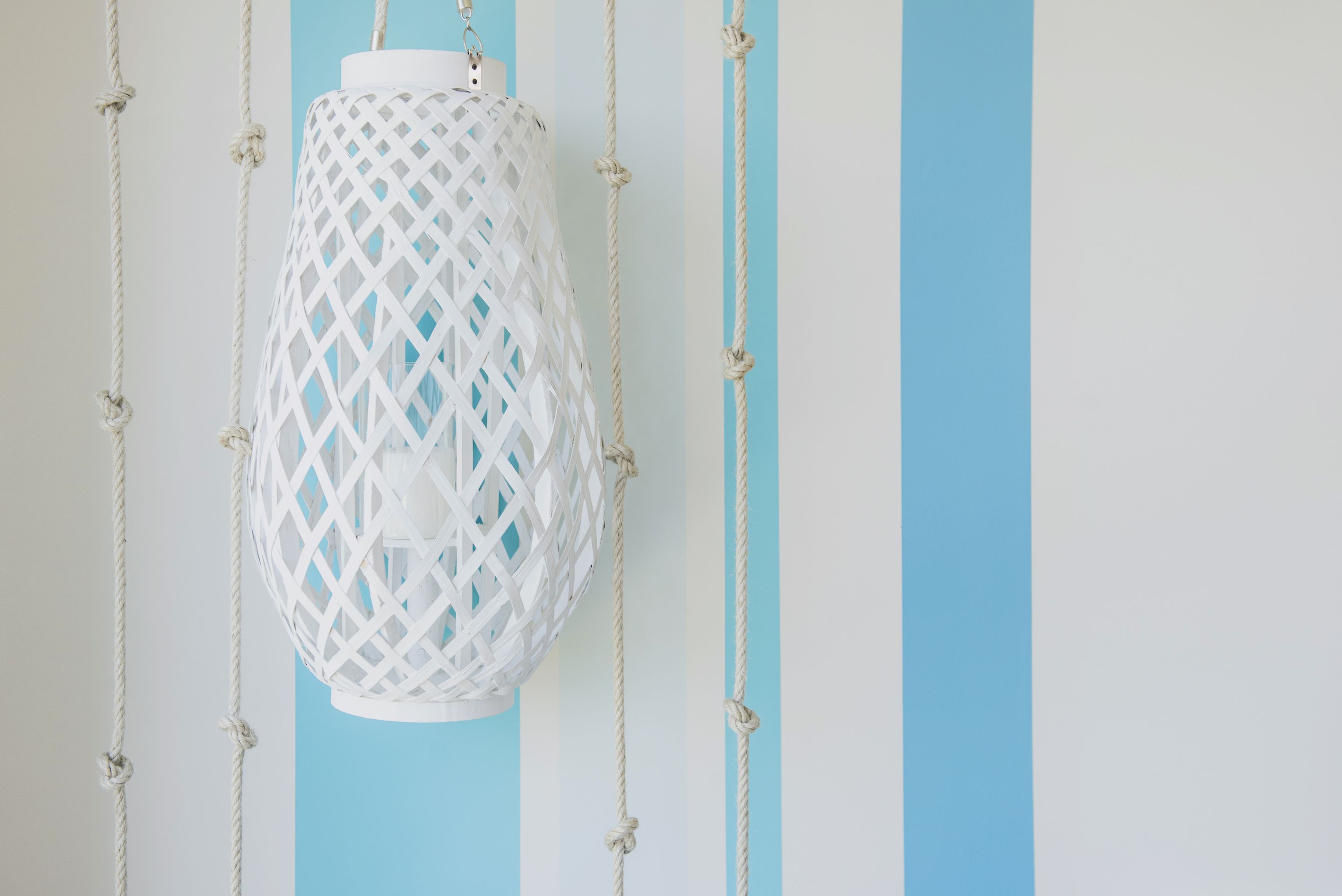 A white hanging lantern with a lattice design, suspended by ropes against a light blue wall.