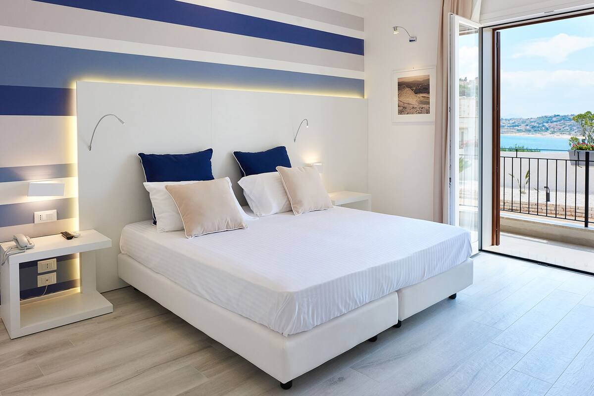 A modern bedroom with a large bed, bedside tables, and a view of the sea.