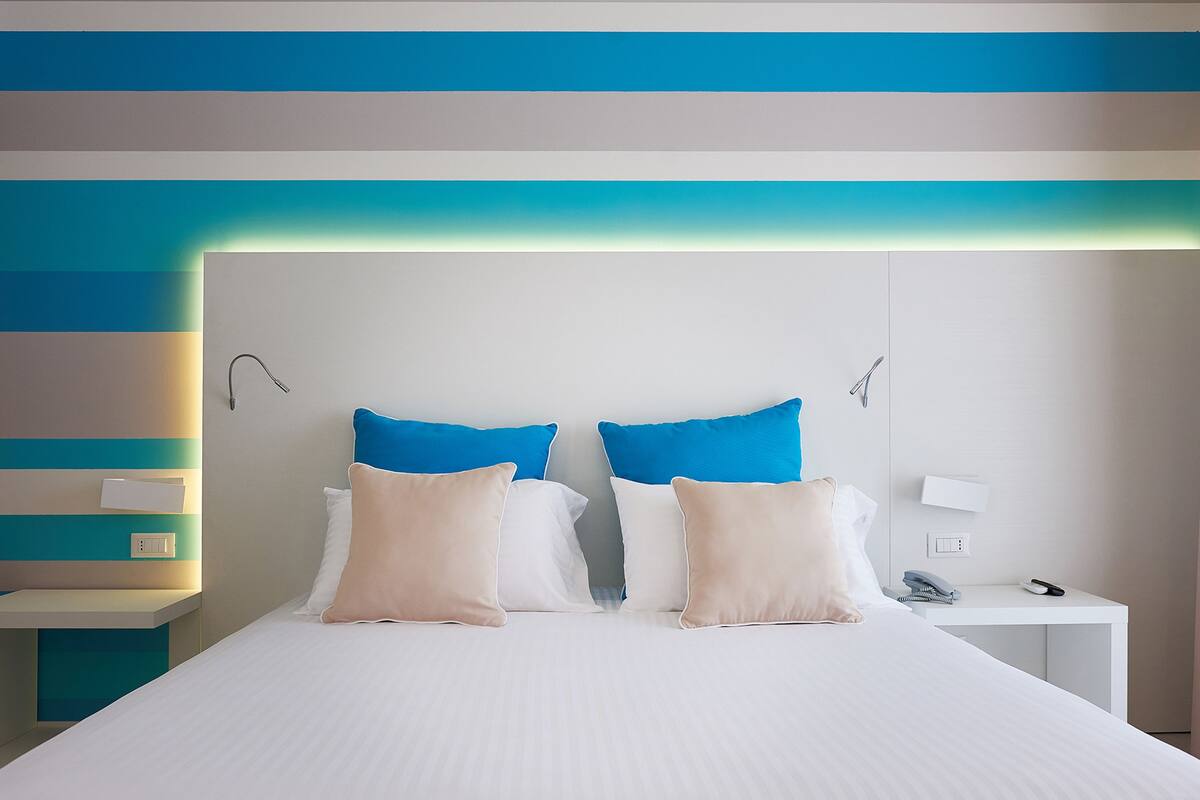 A neatly made bed with white and beige pillows, a white bedside table with a phone and a lamp, and a wall with horizontal stripes in blue and beige.