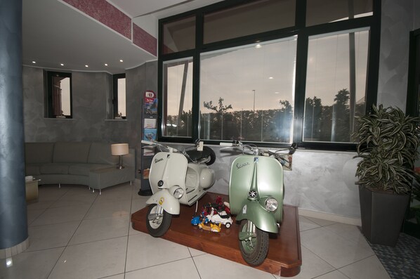 Una sala de estar moderna con un sofá, dos scooters antiguos, una mesita con juguetes y un amplio ventanal con vistas a árboles.