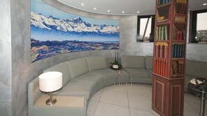 A curved sectional sofa, a tall bookshelf, and a large landscape painting of mountains. - Brindor Hotel (Poirino)