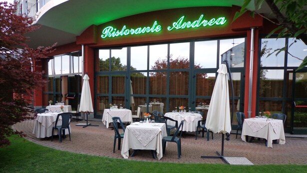An outdoor dining area with tables and chairs, a neon sign reading "Ristorante Andrea," and large windows. - Brindor Hotel (Poirino)
