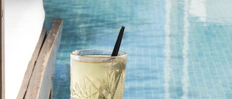 A glass of iced beverage with a straw on a wooden table by a poolside.