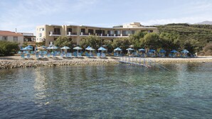 A hotel with clear waters in front, outdoor seating with umbrellas, and a mountainous backdrop. - Almyrida Resort (Apokoronas)