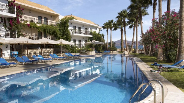 A pool area with lounge chairs and umbrellas, surrounded by palm trees and a building with balconies. - Almyrida Resort (Apokoronas)