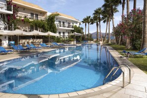A pool area with lounge chairs and umbrellas, surrounded by palm trees and a building with balconies.