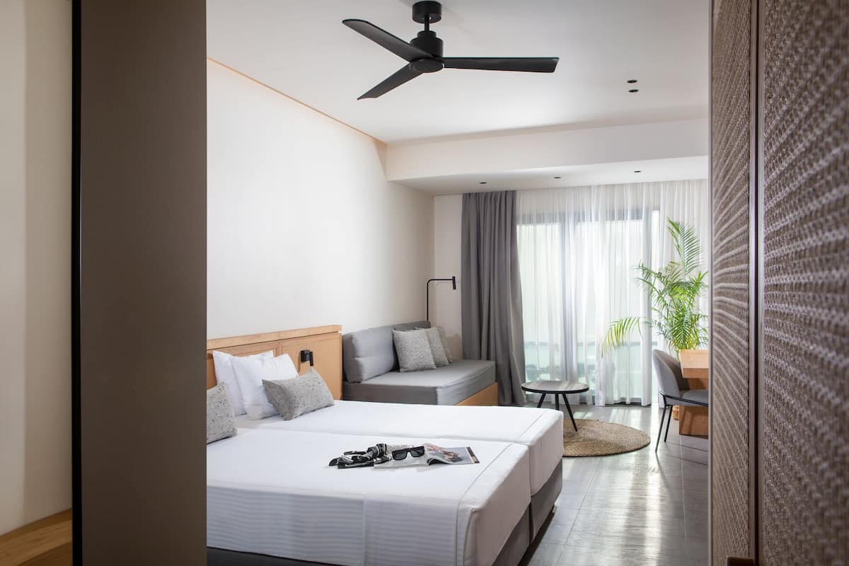 Habitación de hotel moderna con dos camas, ventilador de techo, escritorio y vistas al exterior.