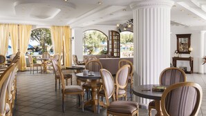 A spacious dining area with round tables and cushioned chairs, large windows with curtains, and a view of the outdoors. - Almyrida Resort (Apokoronas)