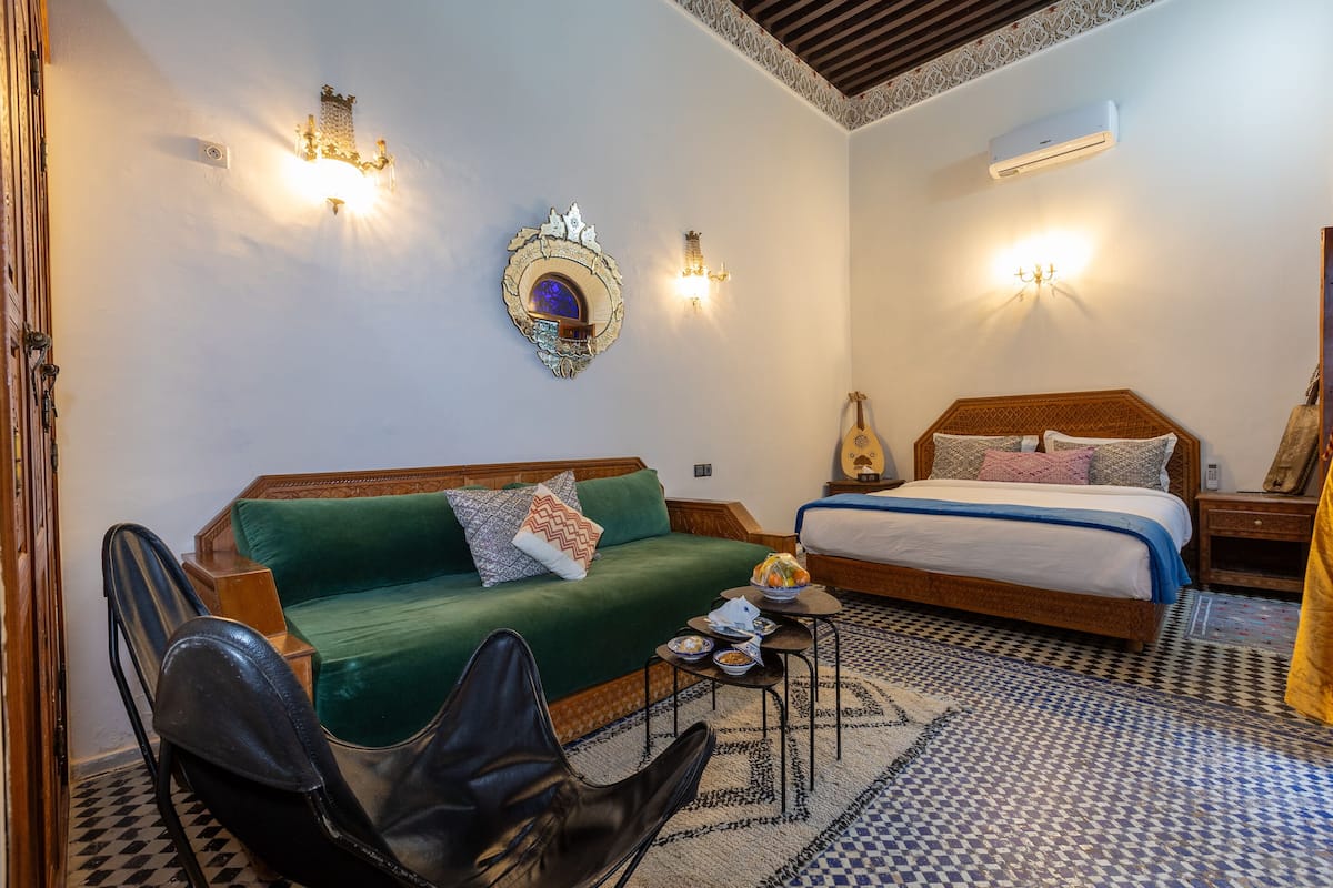 A room with a green sofa, a black leather chair, a wooden bed with a guitar, and a small table with dishes.