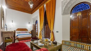 A traditional Moroccan room with a bed, a wooden table, and a red sofa. - Palais Nazha Fes - Luxury Lodging (Fes)