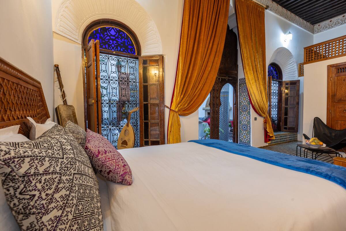 A bedroom with a large bed, ornate wooden headboard, and a view of a courtyard through a blue-tinted door.