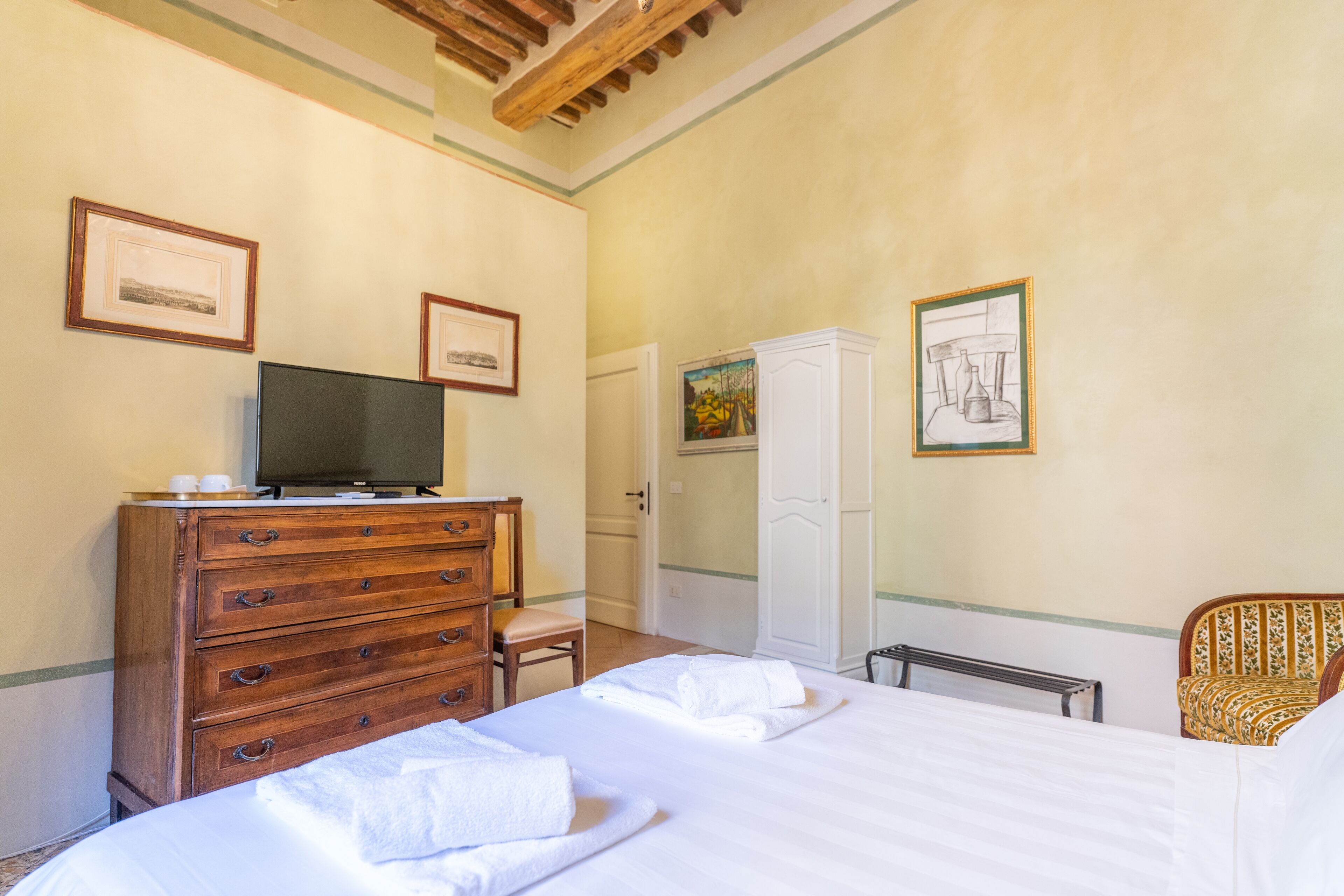 A hotel room with a bed, a wooden dresser, a television, and framed artwork on the walls.