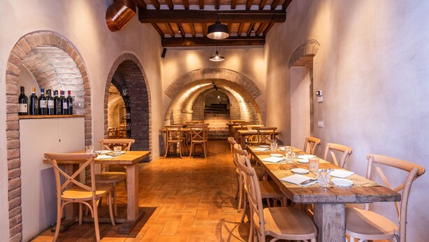 A restaurant with brick arches, wooden tables, and chairs. There are wine bottles on a shelf and a hanging light fixture. - Palazzo Mosela Alla Porta (Montepulciano)
