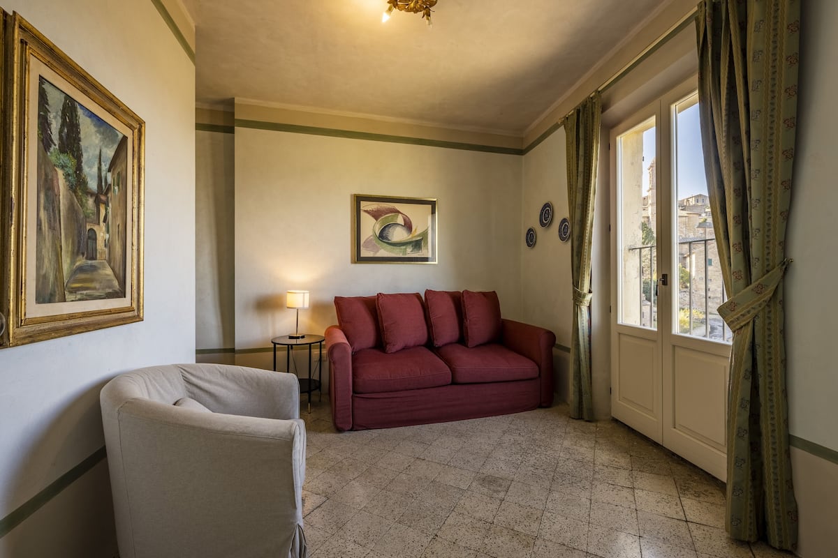 Une pièce avec un canapé rouge, un fauteuil blanc, un tableau, une lampe et une vue sur les immeubles par la fenêtre.