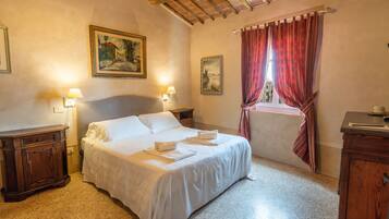 Une chambre d’hôtel comprenant un lit, deux tables de chevet, une petite table, une armoire en bois et une fenêtre avec des rideaux rouges.