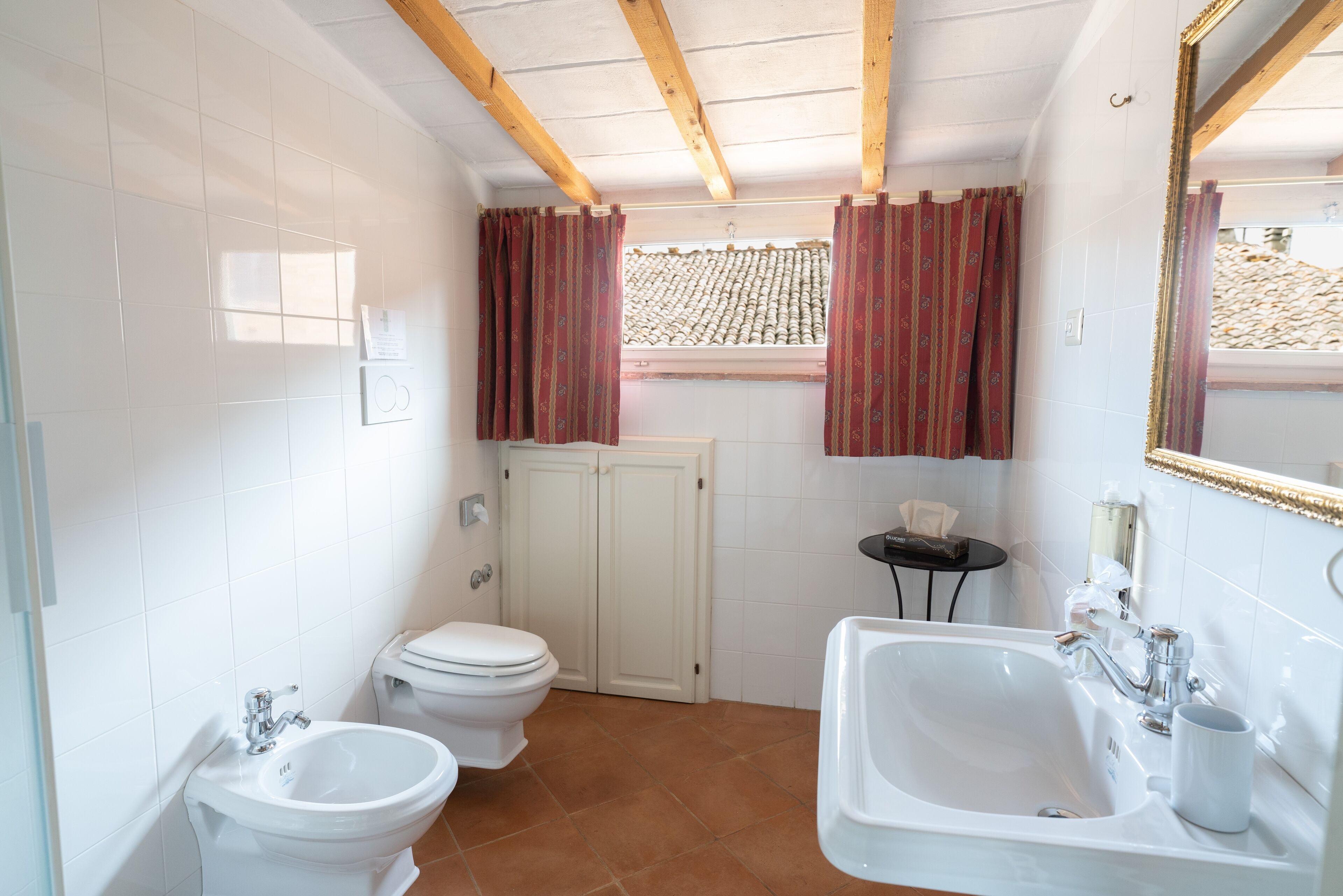 Une salle de bain équipée de toilettes, d’un bidet et d’un lavabo, avec des poutres en bois au plafond et une fenêtre ornée de rideaux rouges.