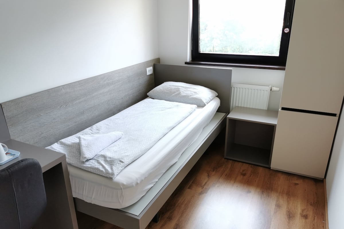 A small, single-bed room with a wooden headboard, a bedside table, a small desk, and a window with a view of greenery.