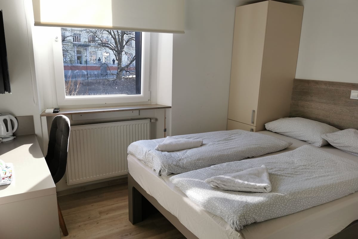 A bedroom with two beds, a desk, a chair, a radiator, and a window with a view of buildings and trees.